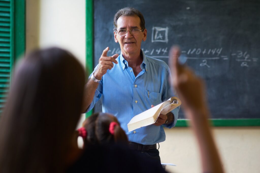 Girl Raising Hand Asking Question To Teacher At School